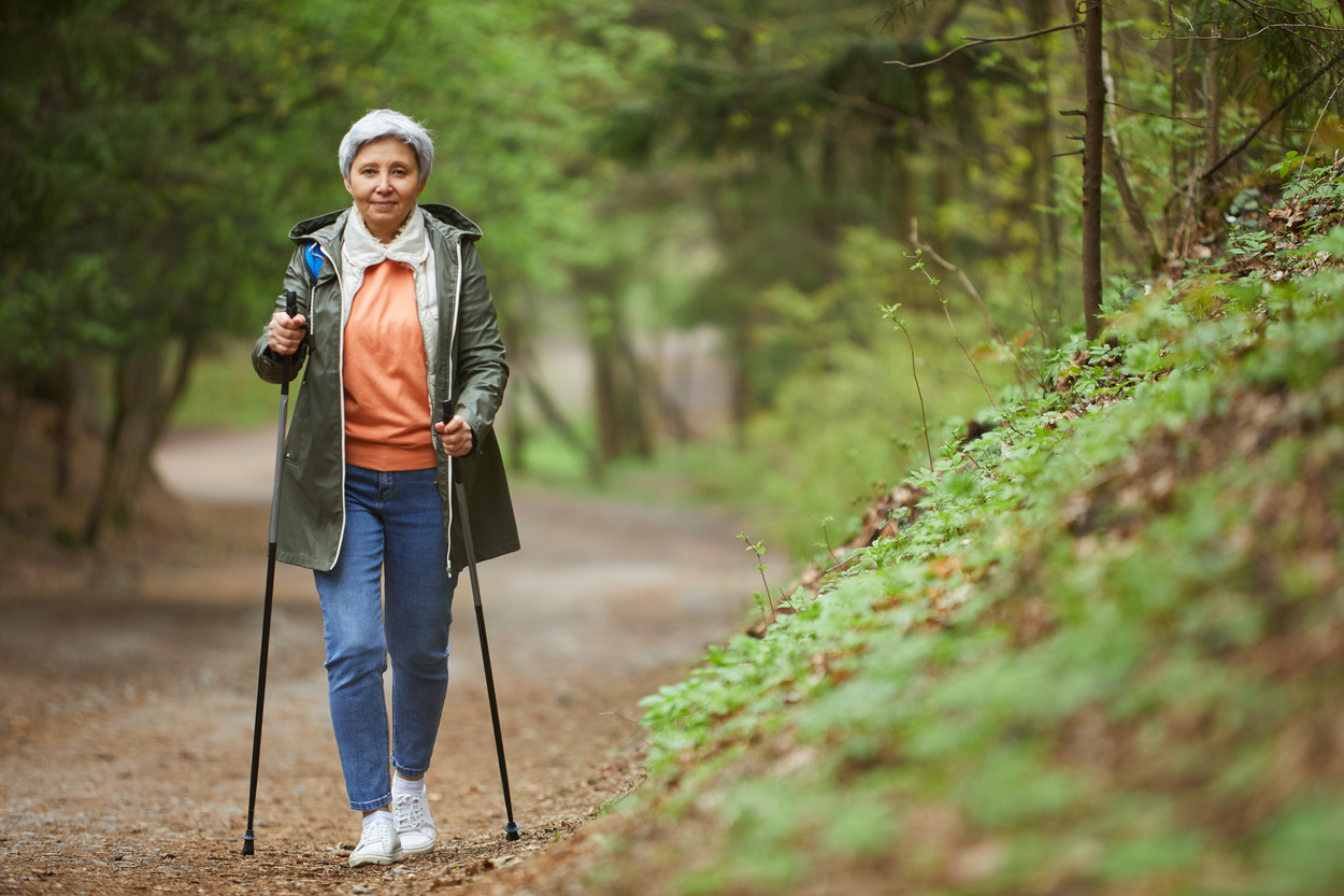 Full length portrait of active senior woman walking towards camera with Nordic poles while enjoying hike in beautiful autumn forest copy space
