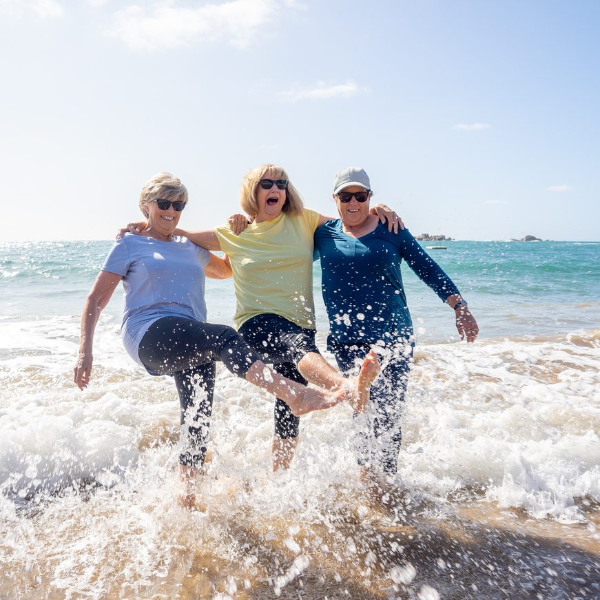 Lovely group of senior girl friends on their 60s walking and having fun splashing water on sea Three mature healthy retired females Laughing and enjoying retirement and outdoors active lifestyle